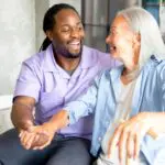 Male nurse in purple top smiles at an elderly female patient while holding her hand