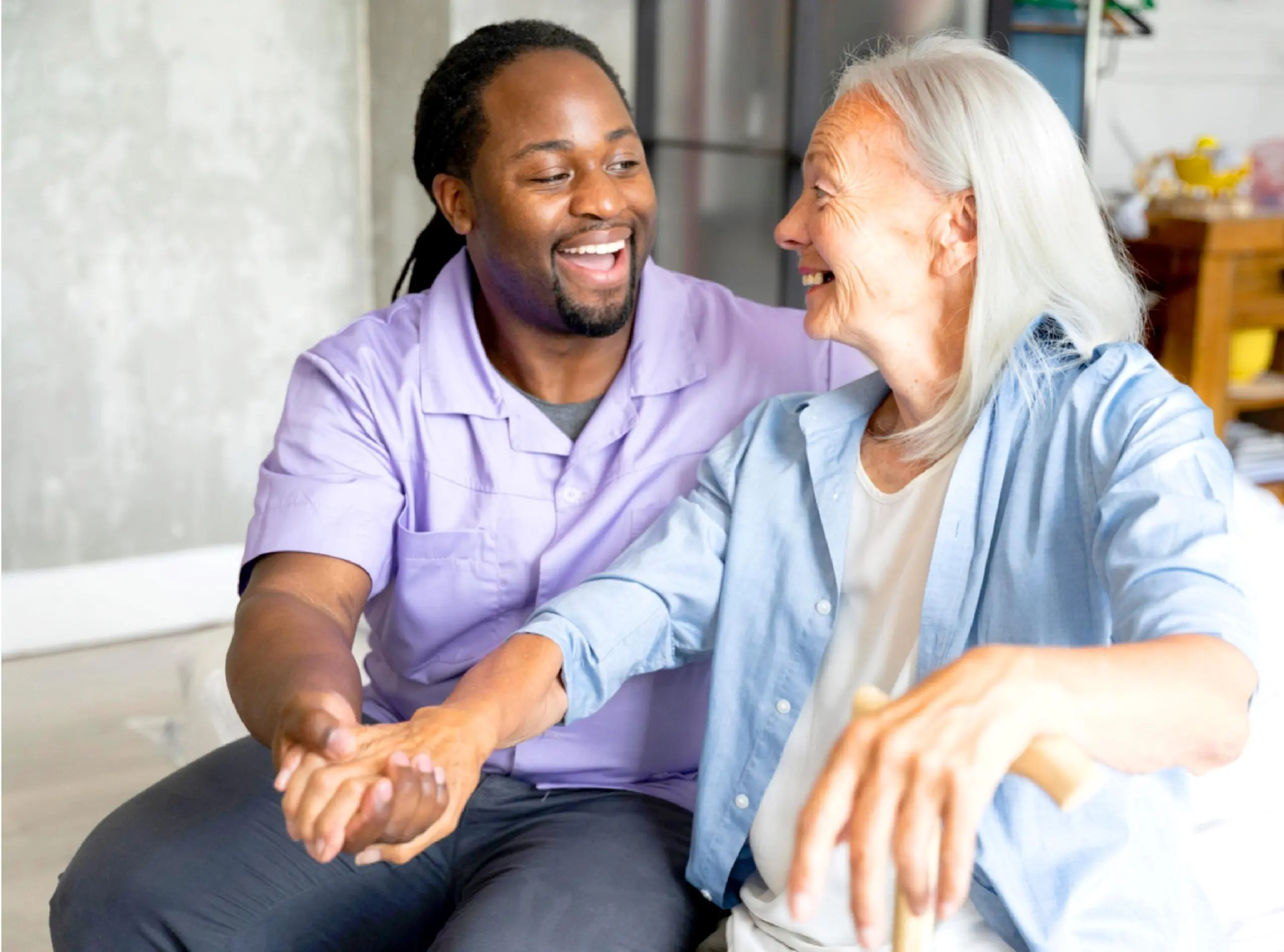 Male nurse in purple top smiles at an elderly female patient while holding her hand