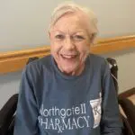 Older woman in a wheelchair and a blue long sleeved tshirt smiles at the camera