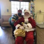 Employee in reindeer antlers and a COVID mask poses with an elderly santa claus in a wheelchair
