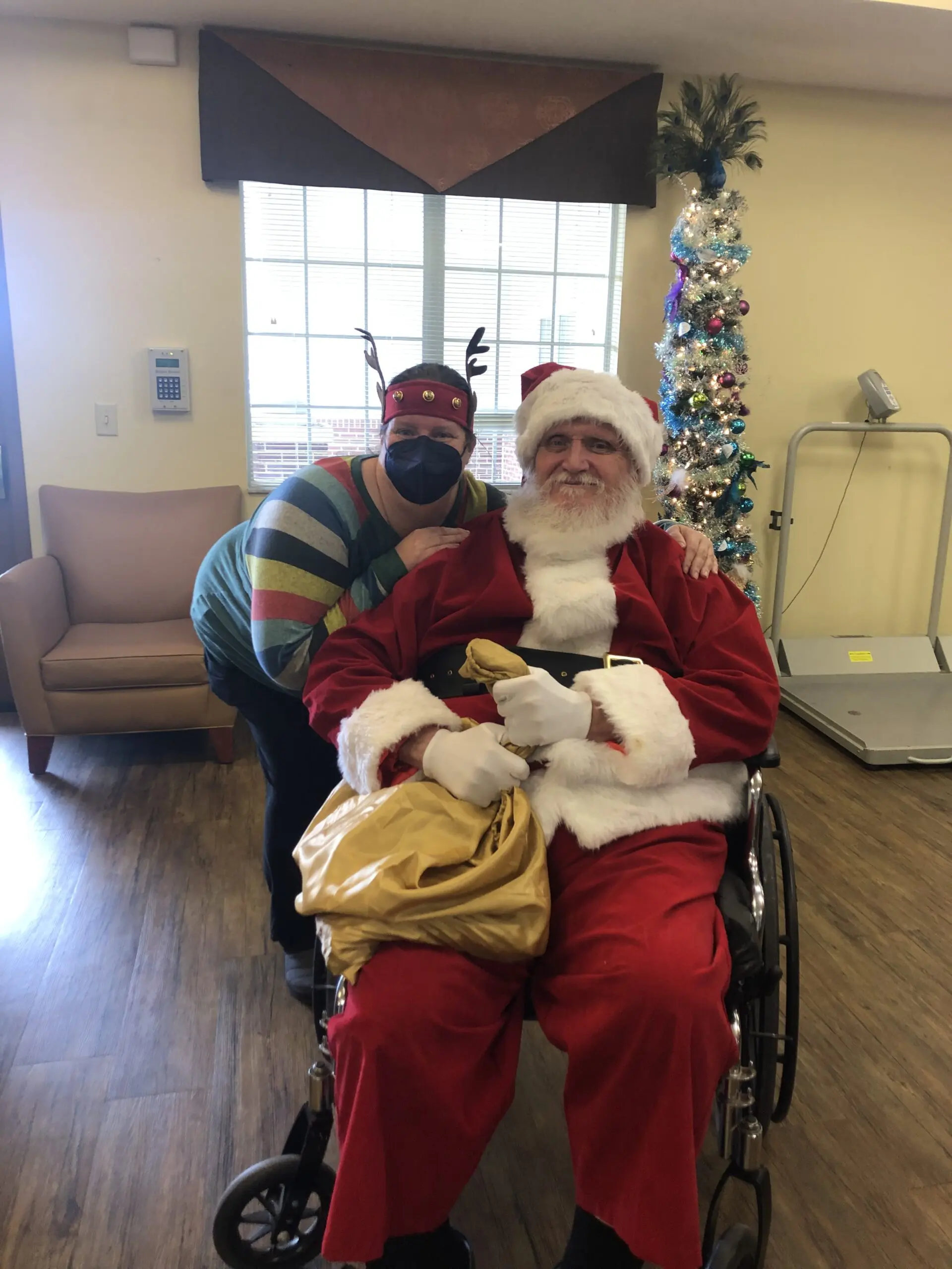 Employee in reindeer antlers and a COVID mask poses with an elderly santa claus in a wheelchair