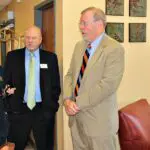 Group of men and women in business clothes stand in a hallway beside a waiting area and talk