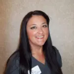 Woman with long dark hair and dark gray scrubs smiles for the camera in front of a beige background