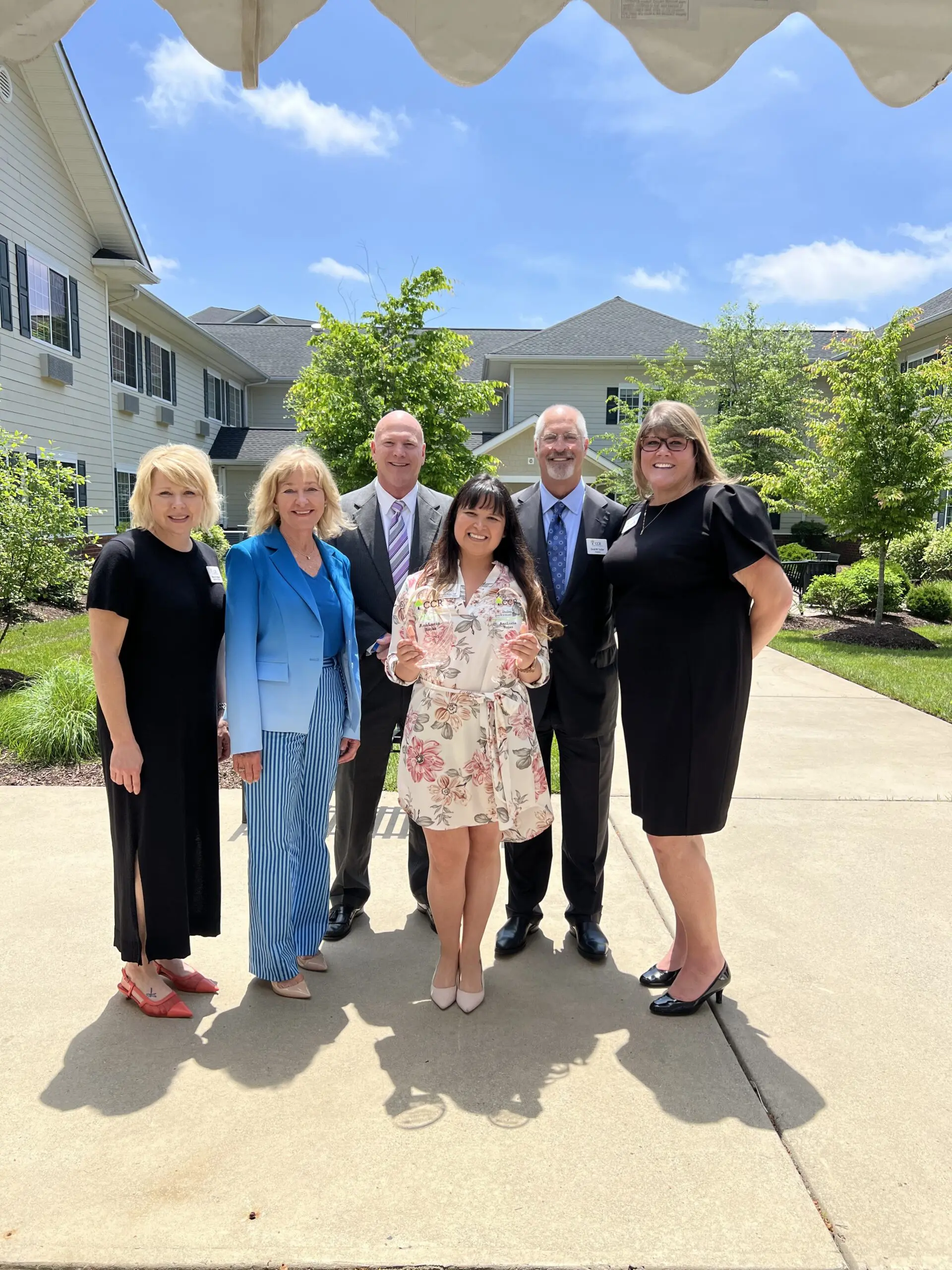 CCR Executives stand in a courtyard with a dark-haired woman in a white floral dress who holds two glass awards