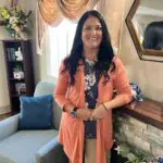Dark-haired woman in pink cardigan and dar blue floral top poses beside a wooden and stone fireplace mantle and smiles for the camera