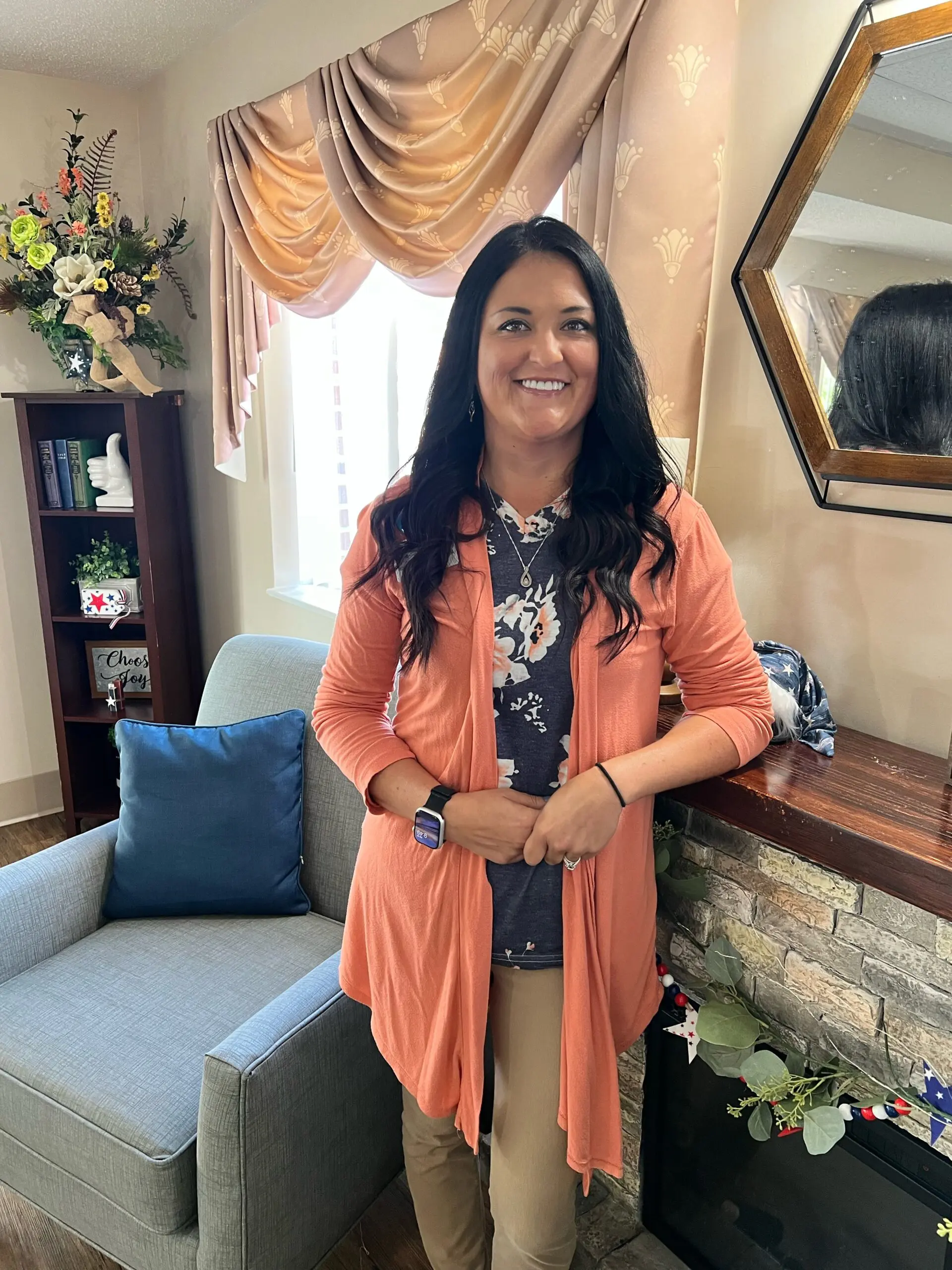 Dark-haired woman in pink cardigan and dar blue floral top poses beside a wooden and stone fireplace mantle and smiles for the camera