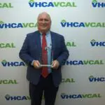 Older man in blue plaid blazer, blue shirt, and red tie holds a glass award while standing in front of a white background with the blue and green VHCAVCAL logo all over it