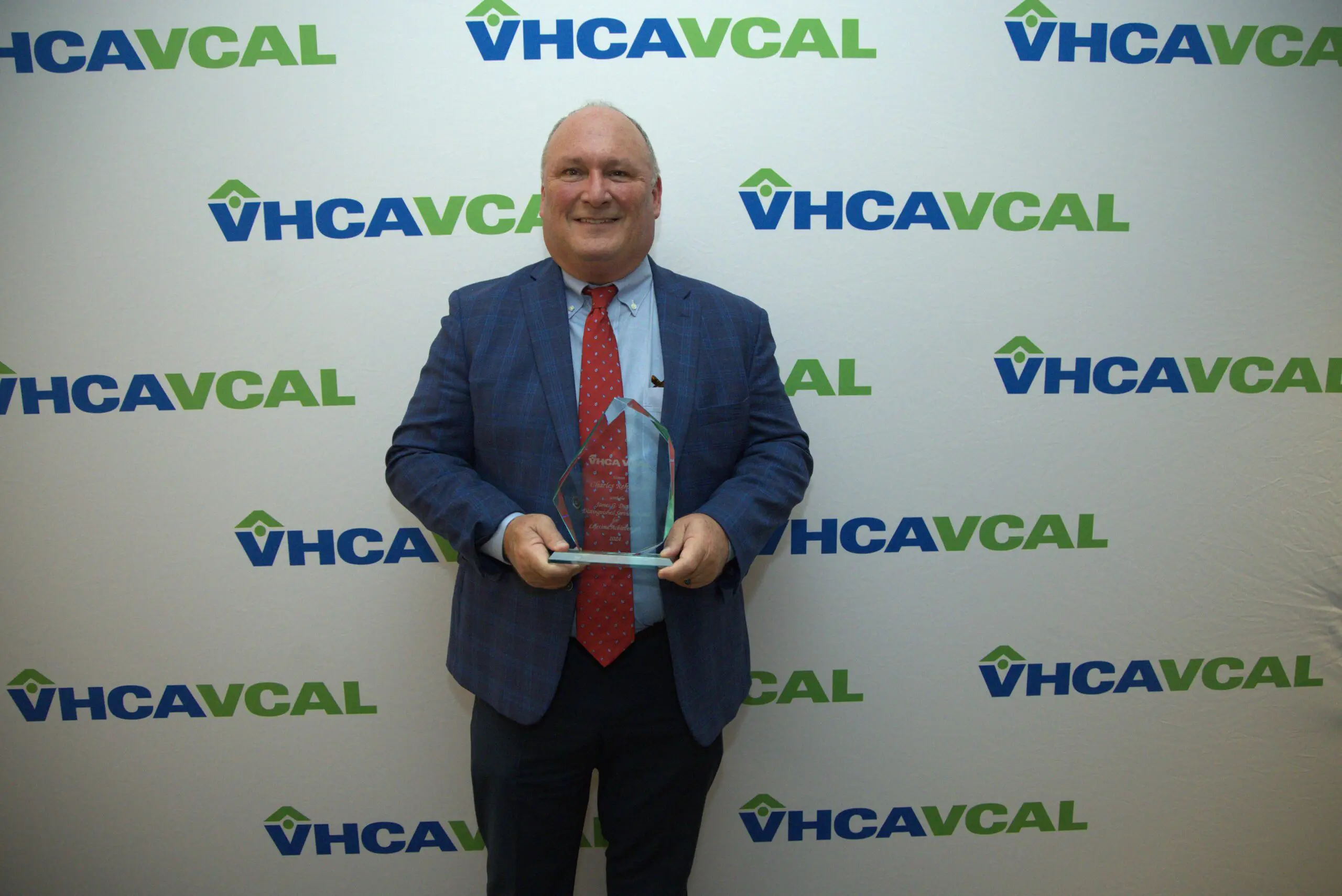 Older man in blue plaid blazer, blue shirt, and red tie holds a glass award while standing in front of a white background with the blue and green VHCAVCAL logo all over it