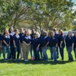 Group of men and women in matching navy blue polos pose together in grass before a row of trees next to a parking lot