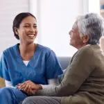 Laughing female nurse in blue scrubs sits on a gray couch with accent pillows, holding the hand of an older woman with short hair and an olive sweater