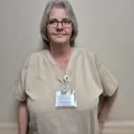Woman with neck-length hair, glasses, and tan scrubs stands in front of a blank neutral wall