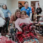 Woman in an orange top smiles with a female patient with an oxygen machine in a wheelchair in a room filled with other patients and caregivers