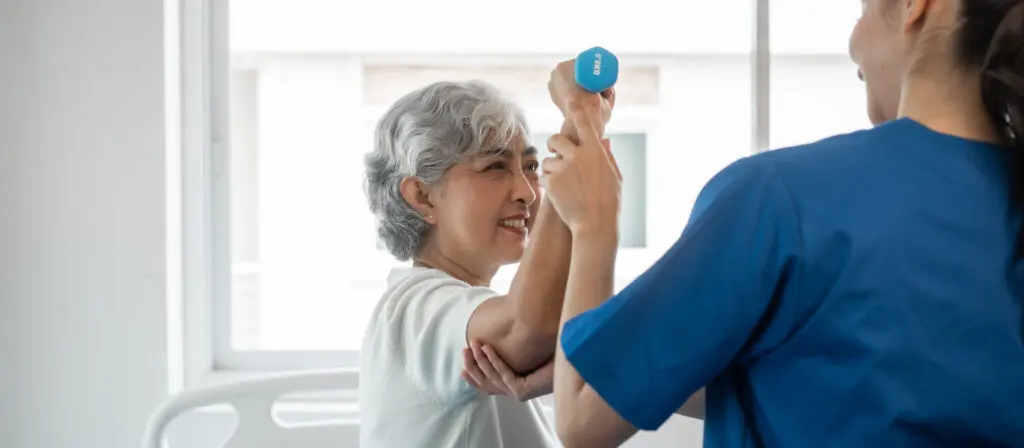 Young physiotherapist helping senior mature asian woman grey hair work out with dumbbells, to recover from injury at health centre in physical therapy session.