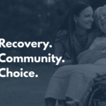 Female nurse in scrubs embraces older woman in a wheelchair, both smiling. White text on a dark overlay reads 