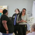 Young woman with long braided hair, glasses, and business clothes laughs with another woman during an event