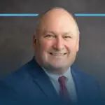 Older bald man in a blue checked suit and red tie smiles in front of a gray blurred background with a blue rectangle around his head