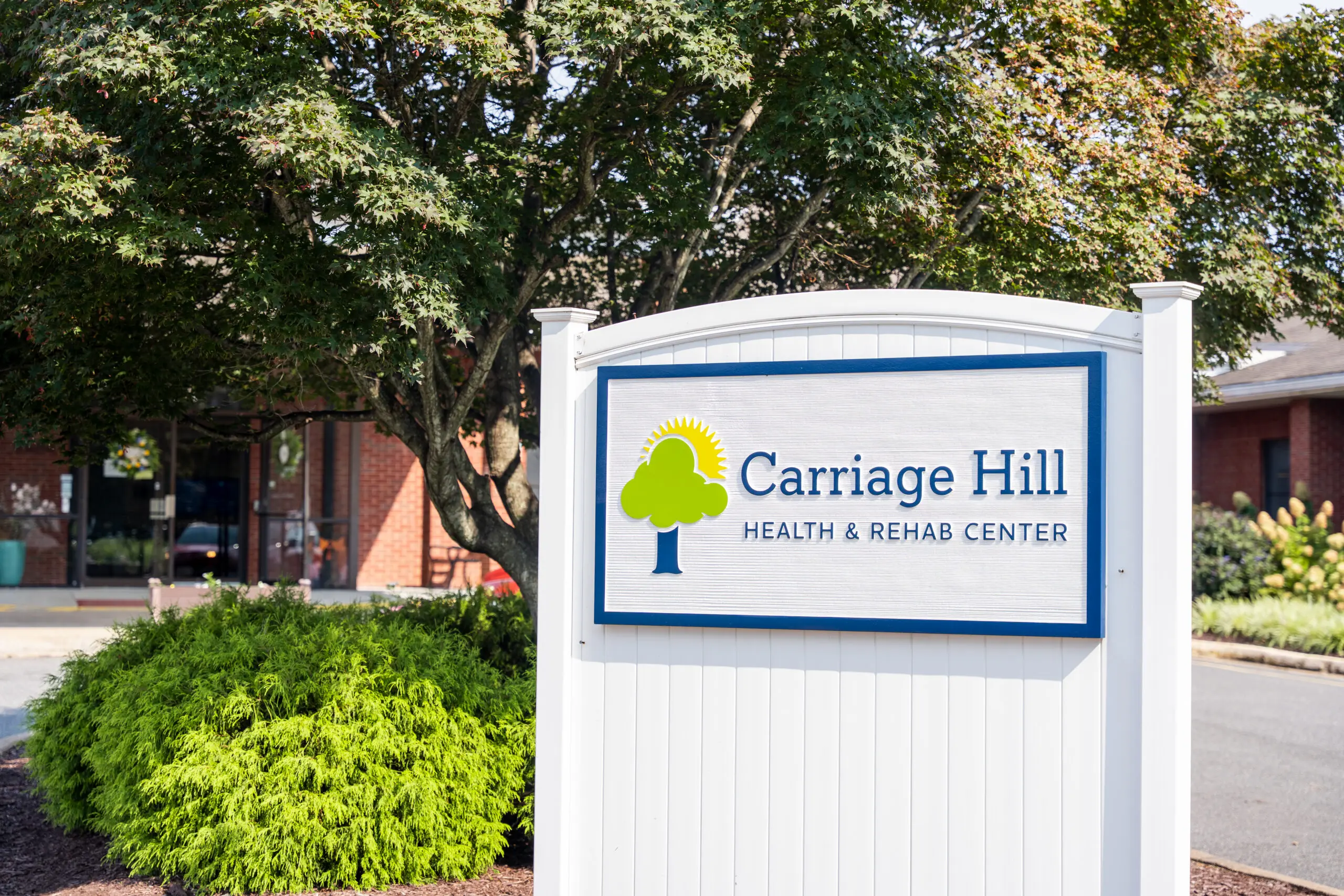 Shot of the Carriage Hill Health & Rehab Center sign, including the Carriage Hill logo mounted on a white fence surrounded by trees and shrubbery.