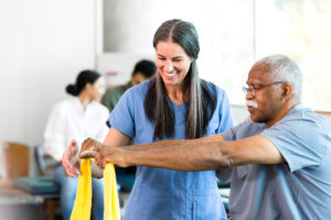 Female nurse in blue scrubs helps an older male patient use a yellow stretching band