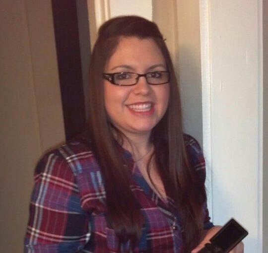 Woman with long dark hair and glasses wears a red plaid flannel and smiles for the camera