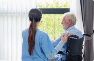 Brunette female nurse with a long ponytail and blue scrubs stands beside an elderly man in a wheelchair in front of a bright window