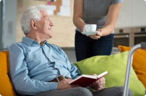 Senior man in blue shirt holds a book and sits on a couch with green and orange pillows while someone hands him a white cup and saucer