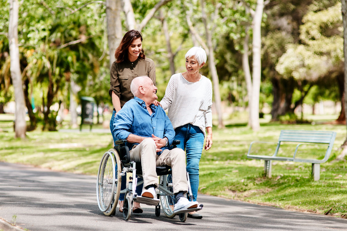 two women and an elderly man in a wheelchair strolling through the park
