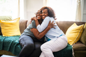 A woman in blue joyfully embracing a senior woman while sitting on a brown suede couch with yellow pillows