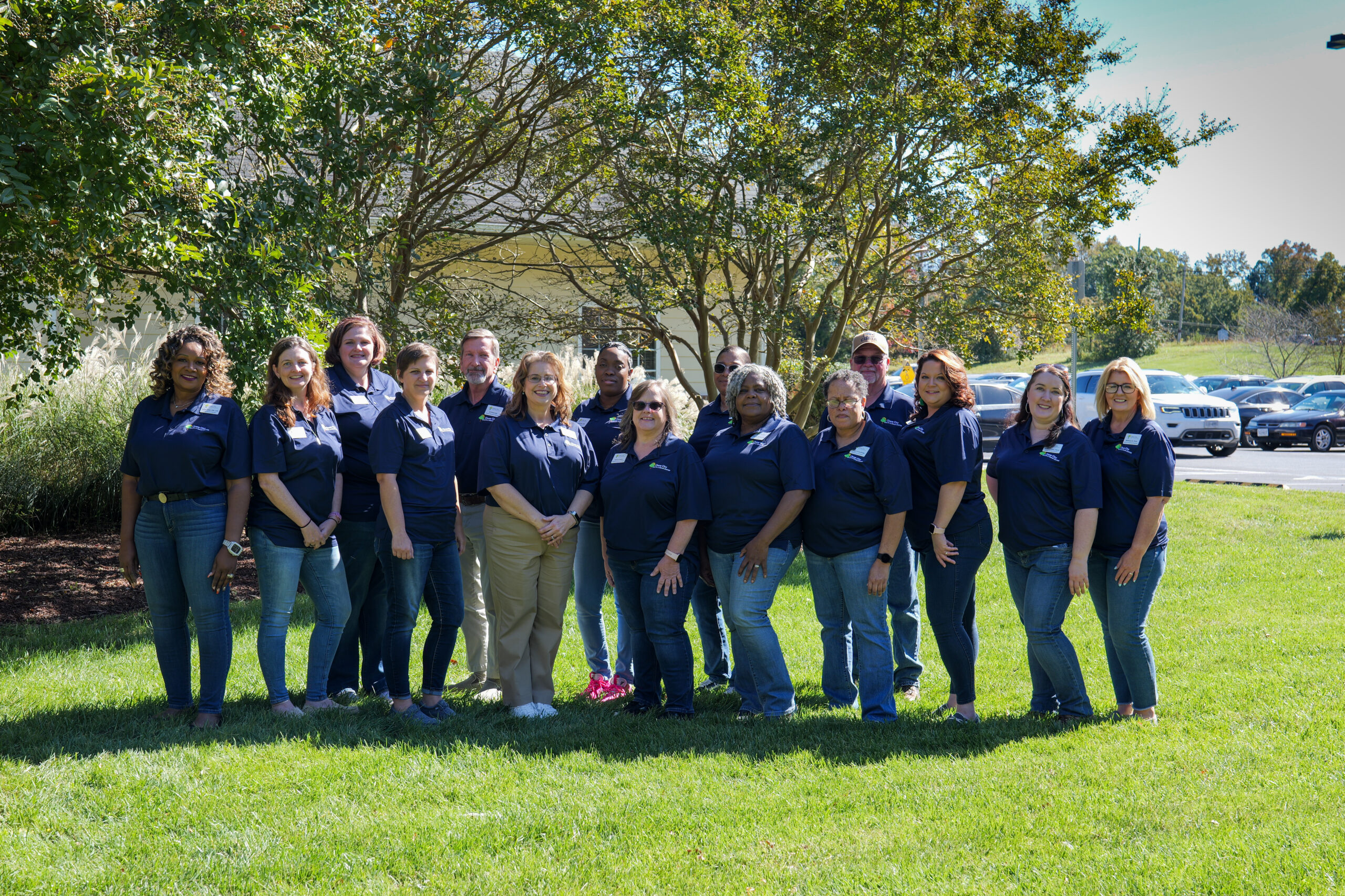 Group of men and women in matching navy blue polo shirts poses for the camera while standing in a grassy area beside a parking lot