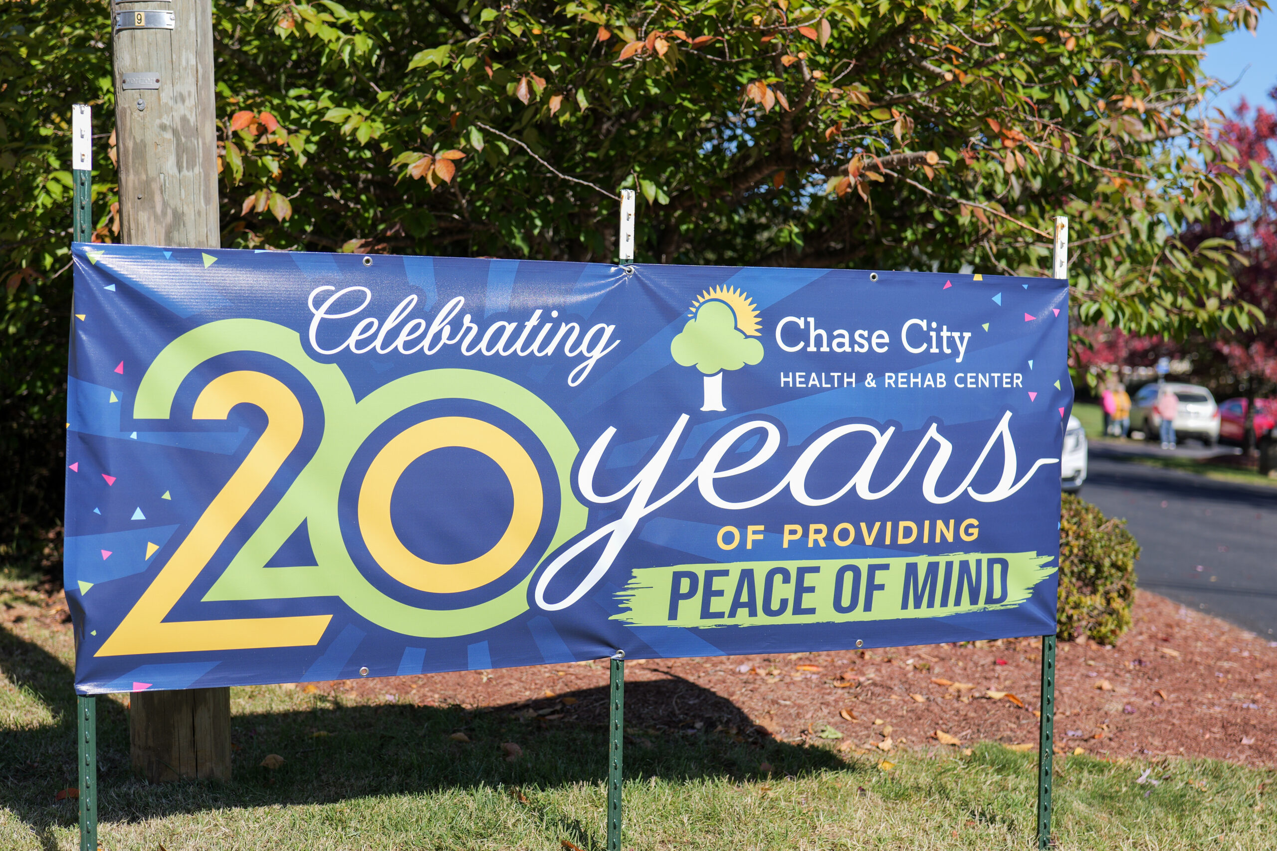 Blue banner in front of green foliage and a telephone pole reads "Chase City Health and Rehab Center, Celebrating 20 Years of Providing Peace of Mind"