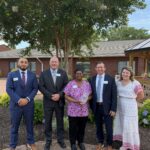Three men in suits, a female nurse in pink scrubs, and a woman in a dress smile together in a garden outside a brick building